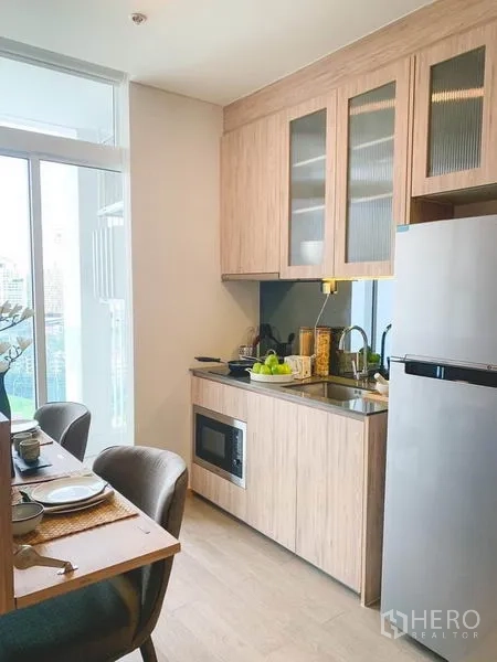 Condominium for sale in Khlong Toei, Bangkok - Efficient kitchenette with light wood cabinets, sink, microwave, and refrigerator beside a window.