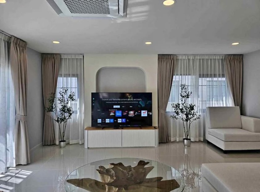 Detached House for rent in Bang Phli, Samut Prakan - Bright living room with TV console, ceiling cassette air‑con, large windows with sheer curtains, potted plants and a glass coffee table.