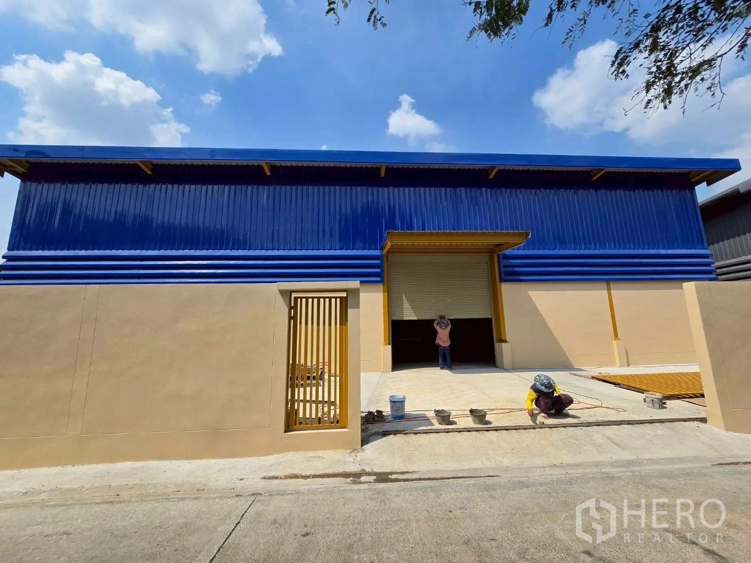 Warehouse for rent in Khlong Sam Wa, Bangkok - Front facade of a modern warehouse with blue panels, beige lower walls, large roller shutter, gate, and loading apron.