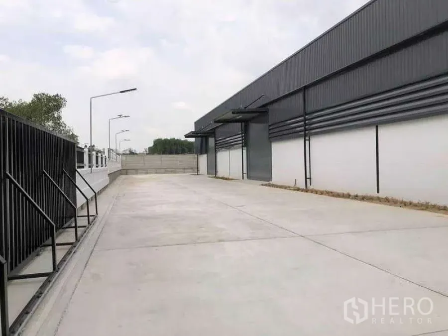 货栈 出租或出售在Nikhom Phatthana, Rayong - 尼空帕他那现代灰色仓库外部，宽阔混凝土装卸庭院、卷帘门与路灯。