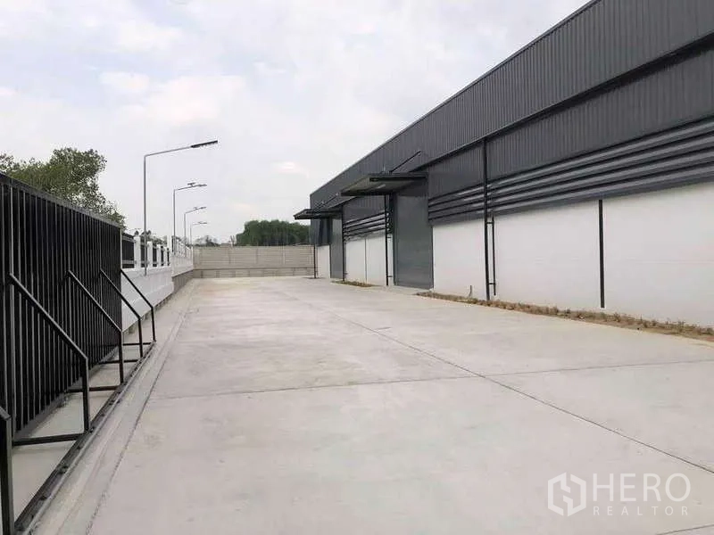 货栈 出租或出售在Nikhom Phatthana, Rayong - 尼空帕他那现代灰色仓库外部，宽阔混凝土装卸庭院、卷帘门与路灯。