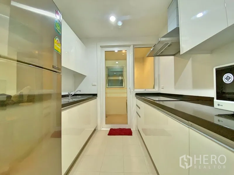 Condominium for rent in Khlong Toei, Bangkok - Enclosed galley kitchen with white cabinetry, black countertops and stainless refrigerator.