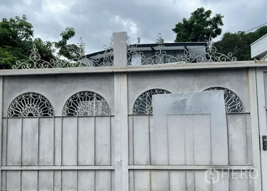 Detached House for rent in Phra Khanong, Bangkok - Two-storey house roofline peeking over an ornate boundary wall with wrought-iron details and greenery.