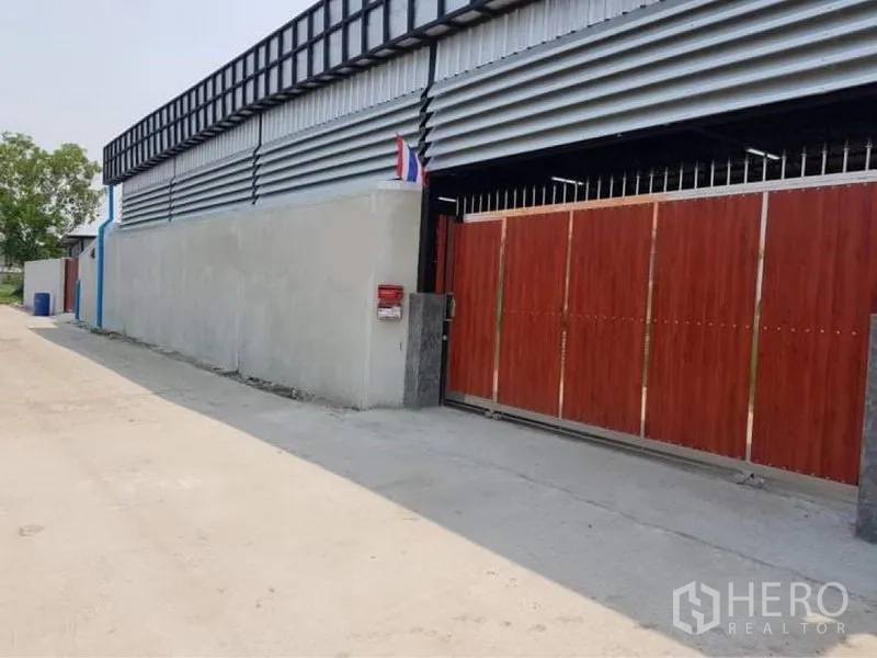 Warehouse for sale in Lam Luk Ka, Pathum Thani - Street-side view of a secure metal-clad warehouse with large wooden sliding gate in Lam Luk Ka, Pathum Thani.