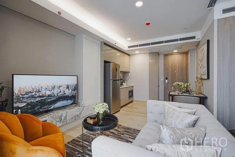 Condominium for sale in Khlong Toei, Bangkok - Bright living room with TV on a marble console, sofa seating and a linear kitchenette along the wall.