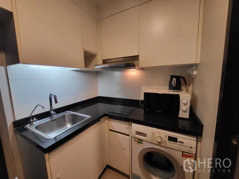 Condominium for rent or sale in Phaya Thai, Bangkok - Compact L-shaped kitchen with black countertops, sink, cooktop, microwave and washing machine.