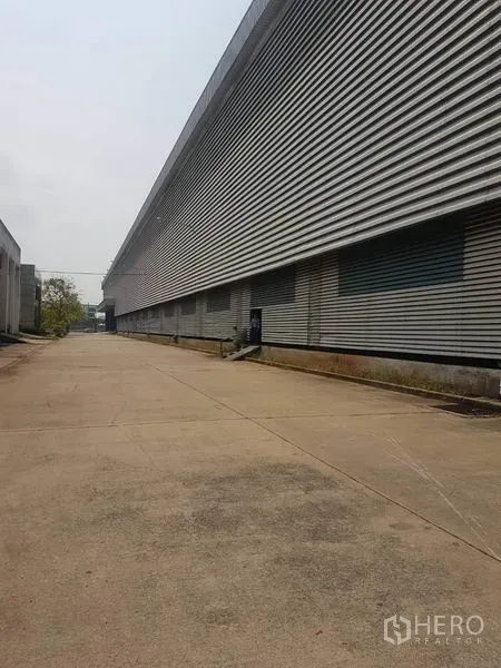 Factory for rent in Mueang Samut Sakhon, Samut Sakhon - Wide concrete driveway beside a long corrugated‑metal factory exterior with multiple loading bays in Samut Sakhon.