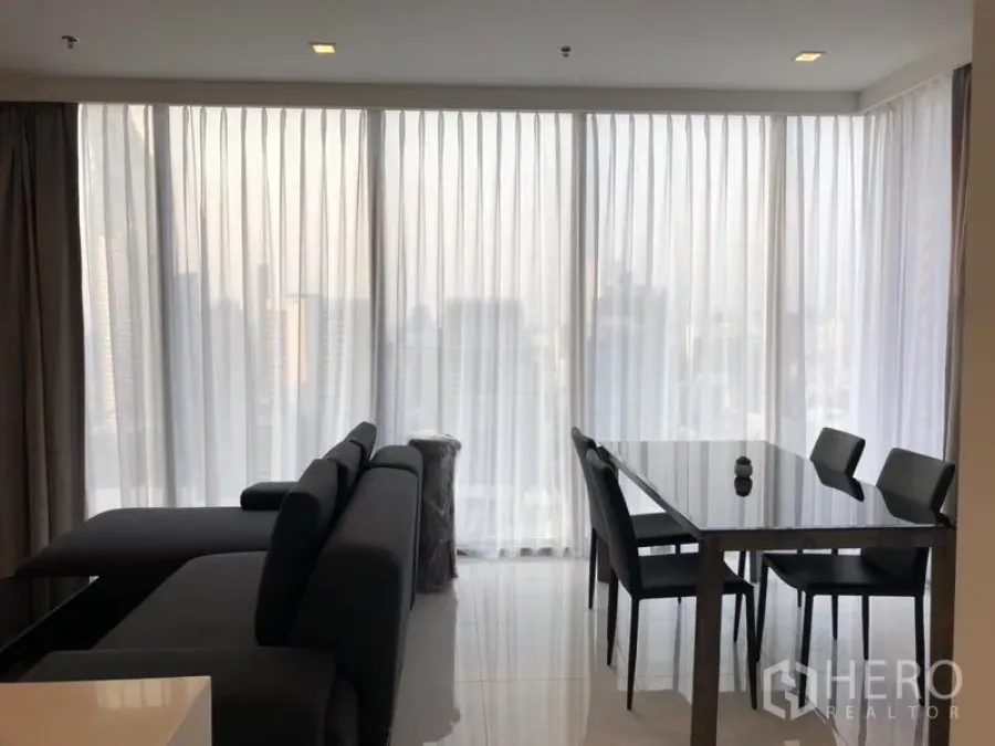 Condominium for rent in Sathon, Bangkok - Bright living and dining area with floor‑to‑ceiling sheer curtains, city skyline views, a dark sectional sofa and a glass dining table.