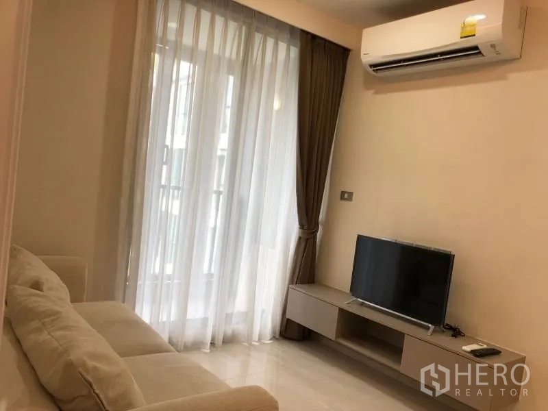 Condominium for sale in Khlong Toei, Bangkok - Living room with sofa, TV console, air‑conditioner and balcony doors with sheer curtains.