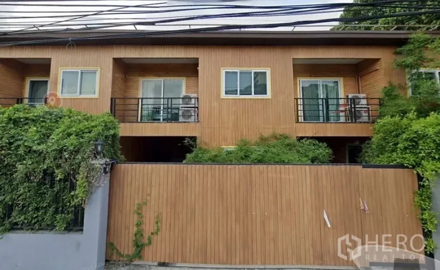 ทาวน์เฮาส์ ให้เช่าหรือขายในWatthana, Bangkok - ด้านหน้าทาวน์เฮาส์สองชั้นสไตล์โมเดิร์น ผนังลายไม้ ระเบียงคู่ ประตูกระจกบานเลื่อน ราวเหล็กสีดำ พร้อมกำแพงรั้วและไม้พุ่มเขียวชอุ่มในย่านสุขุมวิท–เอกมัย