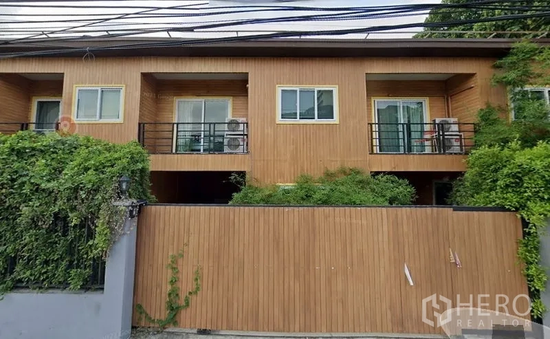 ทาวน์เฮาส์ ให้เช่าหรือขายในWatthana, Bangkok - ด้านหน้าทาวน์เฮาส์สองชั้นสไตล์โมเดิร์น ผนังลายไม้ ระเบียงคู่ ประตูกระจกบานเลื่อน ราวเหล็กสีดำ พร้อมกำแพงรั้วและไม้พุ่มเขียวชอุ่มในย่านสุขุมวิท–เอกมัย