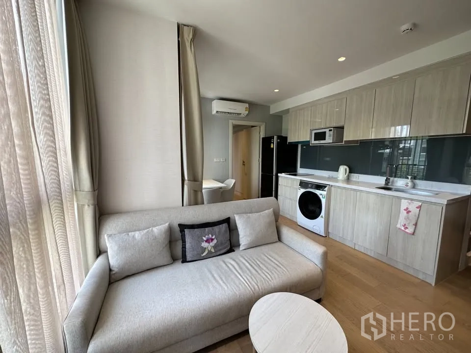 คอนโด for rent in Watthana, Bangkok - พื้นที่นั่งเล่นแบบโอเพ่นพร้อมโซฟาและครัวยาวมีตู้เก็บของ ไมโครเวฟ ตู้เย็น และเครื่องซักผ้า.