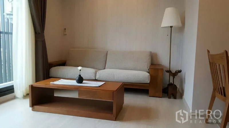 Condominium for rent in Khlong Toei, Bangkok - Bright living room with beige sofa, wooden coffee table and floor lamp beside the balcony window.