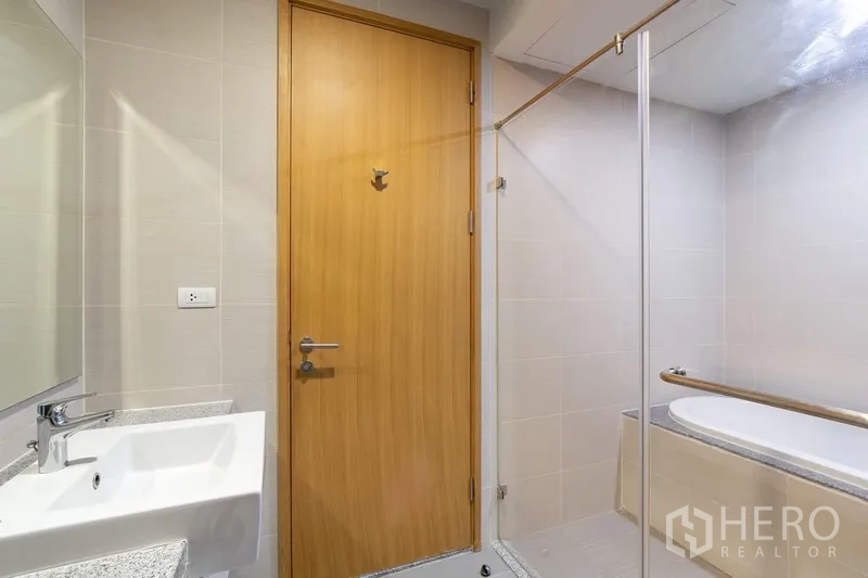 Condominium for rent in Khlong Toei, Bangkok - Contemporary bathroom with glass shower screen, bathtub, and wooden door.