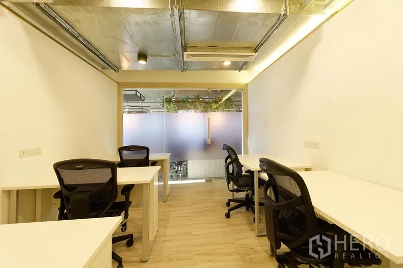 Office Space for rent in Watthana, Bangkok - 14 sqm workspace with ergonomic chairs, LED lighting, exposed ductwork ceiling and a frosted glass door facing greenery.