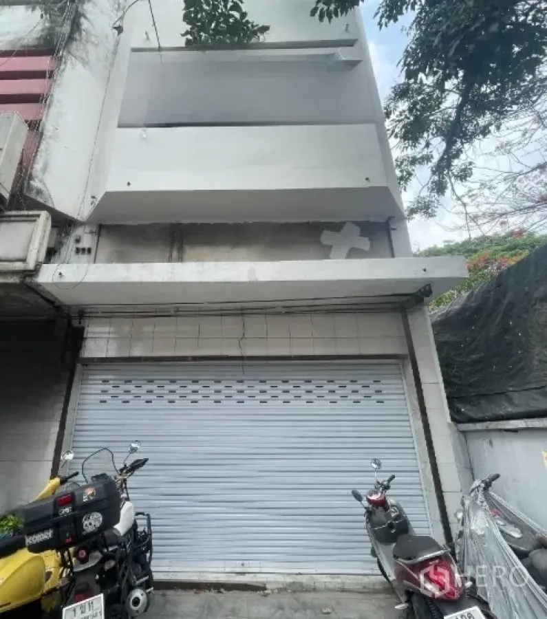 店屋 出租在Watthana, Bangkok - 白色多层店屋外立面，金属卷闸门关闭，前方停放机车。