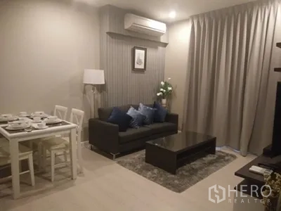 คอนโด ให้เช่าในHuai Khwang, Bangkok - ห้องนั่งเล่นอบอุ่นพร้อมโซฟาสีเทา โต๊ะกลาง พรม และมุมรับประทานอาหารใต้แสงไฟนุ่ม