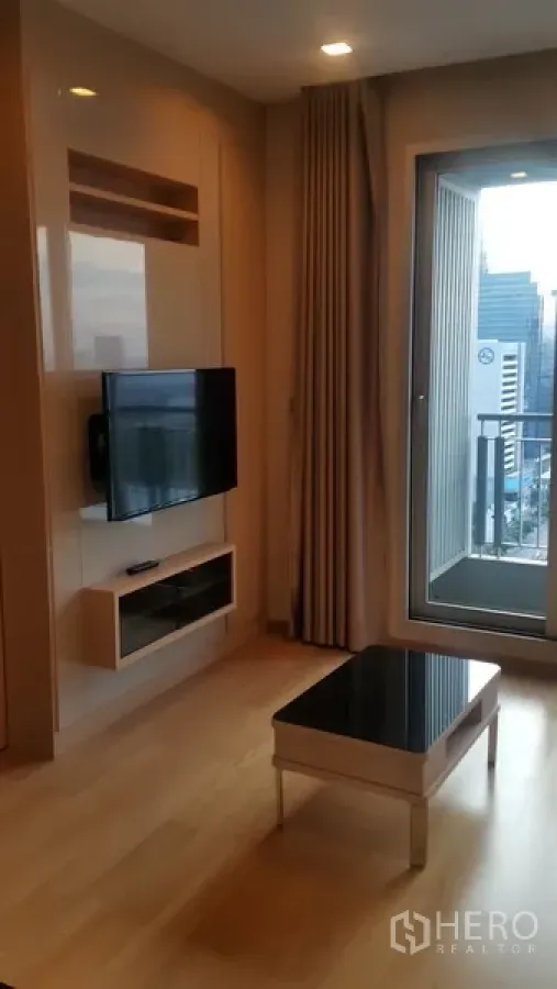 公寓 出租在Ratchathewi, Bangkok - 明亮客厅，墙挂电视、木地板，落地门通向阳台，可观曼谷天际线。