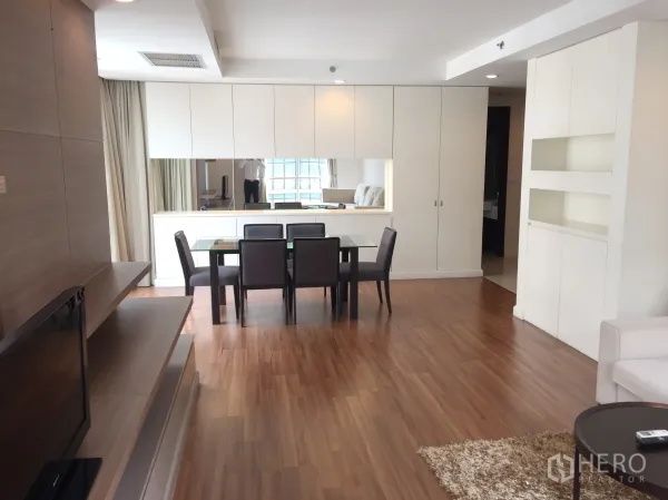 Condominium for rent in Pathum Wan, Bangkok - Bright open-plan living and dining area with wood flooring, glass dining table and built-in white cabinetry with a large mirror.