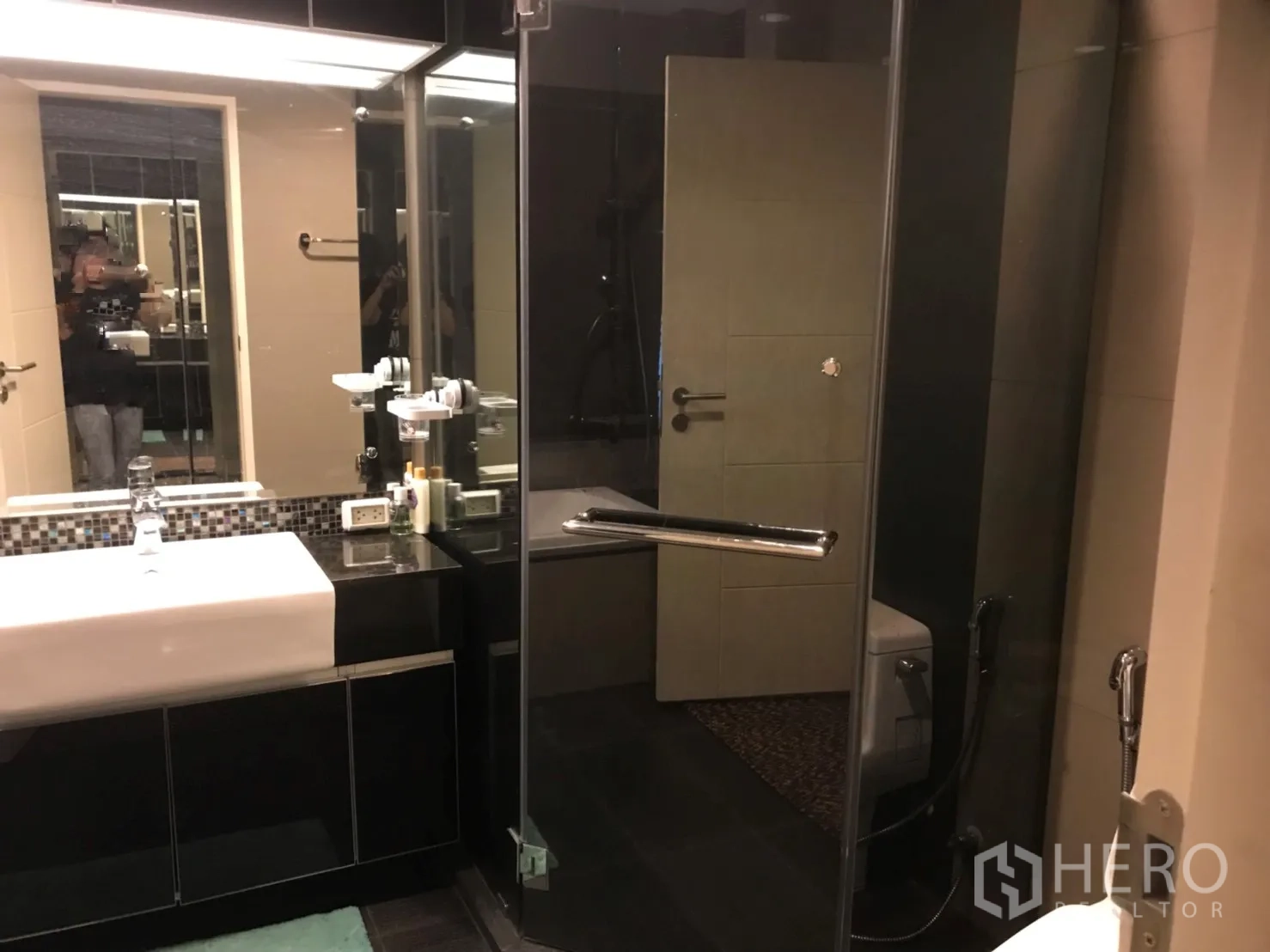 Condominium for rent in Khlong Toei, Bangkok - Modern bathroom with a glass-enclosed shower, large mirror and sleek dark vanity.