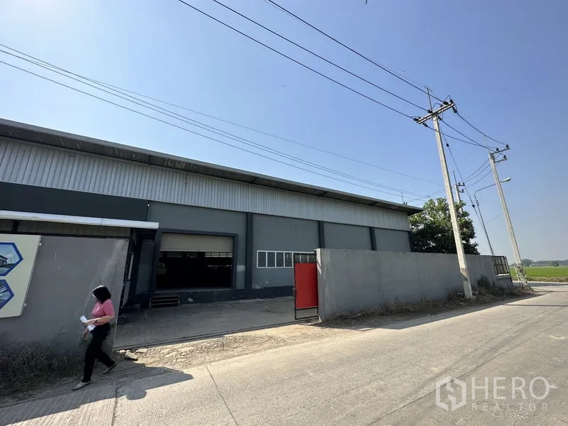 Factory for rent in Lam Luk Ka, Pathum Thani - Street view of a grey factory warehouse with roller shutter door, red gate and perimeter wall.