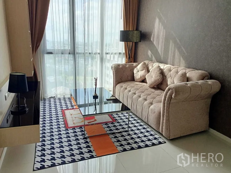 公寓 出租在Watthana, Bangkok - 客厅明亮，米色纽扣沙发、落地窗与薄纱窗帘，配玻璃茶几。