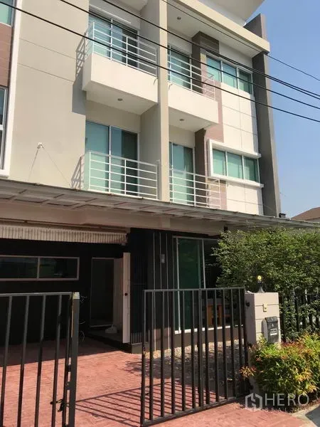 Townhouse for rent or sale in Suan Luang, Bangkok - Modern three-storey townhouse exterior with balconies, gated driveway and shrubs.