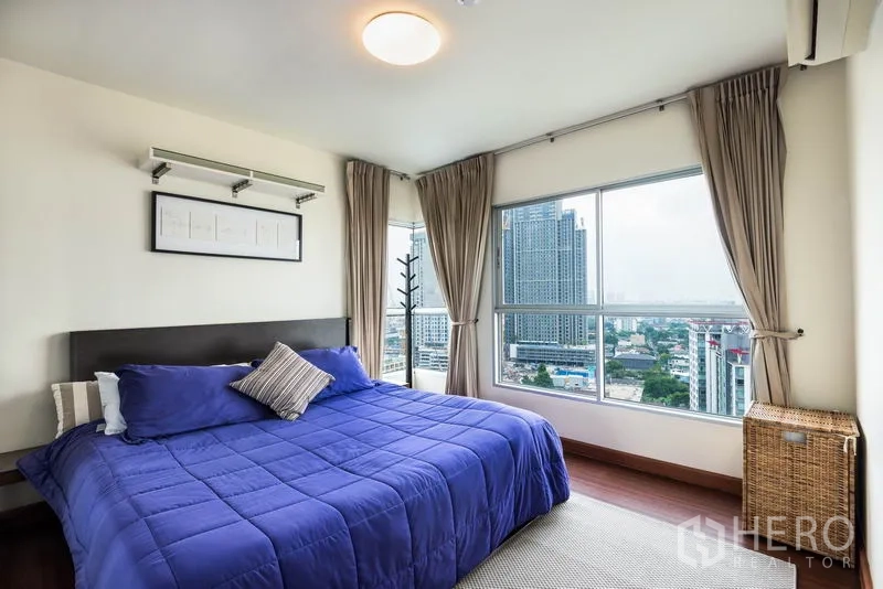 Condominium for rent in Bang Na, Bangkok - Corner master bedroom with blue bedding and panoramic skyline windows.