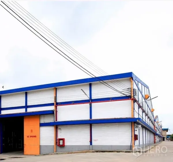 Factory for rent in Samut Prakan - Exterior of a modern factory in Phra Pradaeng with blue-and-white facade, orange loading doors, and a wide driveway.
