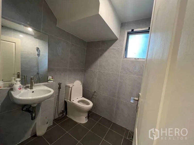 Detached House for sale in Bang Kruai, Nonthaburi - Ground‑floor bathroom with grey tiles, window, wall‑mounted sink and toilet.