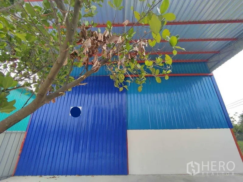 货栈 出租在Pak Kret, Nonthaburi - 外立面特写：蓝色金属板墙面，前方绿树遮阴。