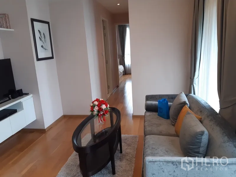 公寓 出租在Watthana, Bangkok - 温馨客厅，灰色沙发、电视柜与通往卧室的走廊，木地板与窗帘。