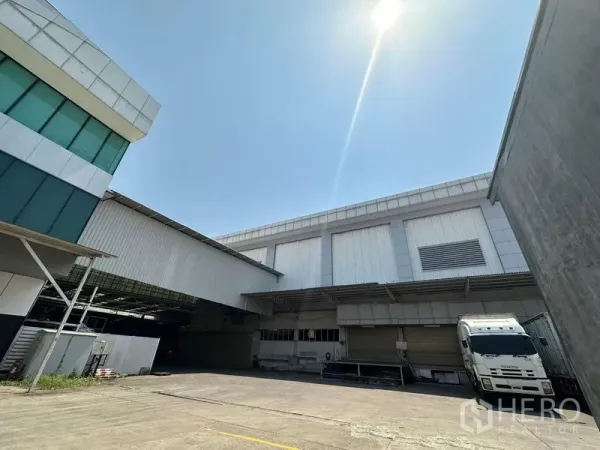 Warehouse for rent in Lam Luk Ka, Pathum Thani - Modern warehouse exterior with covered loading bays and a parked truck under a bright sky.