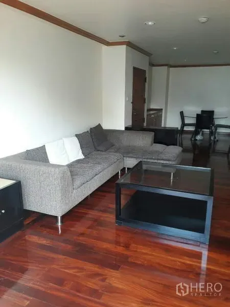 公寓 出租在Pathum Wan, Bangkok - 客厅一角，灰色L型沙发搭配玻璃茶几与亮泽木地板。