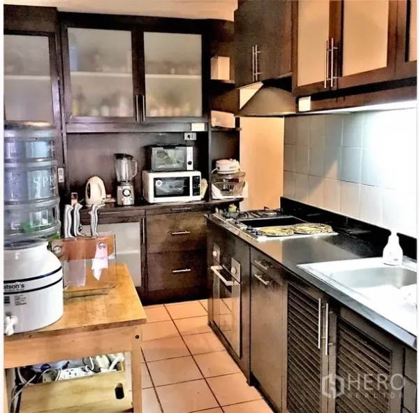 Condominium for rent or sale in Watthana, Bangkok - Separate kitchen with dark wood cabinets, gas hob, oven, microwave, and tiled flooring.