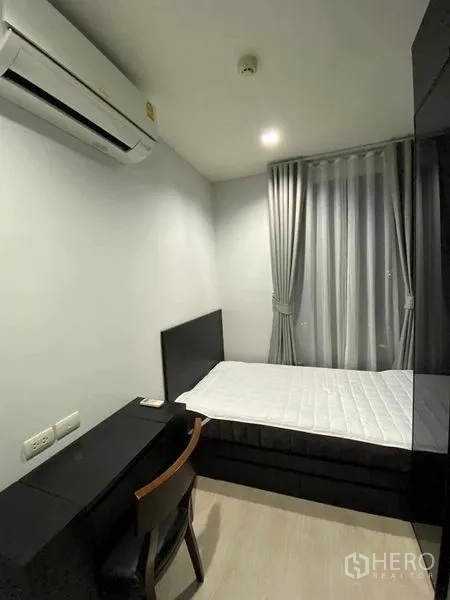 Condominium for rent in Khlong Toei, Bangkok - Second bedroom with single bed, study desk, grey curtains and wall-mounted air conditioner.