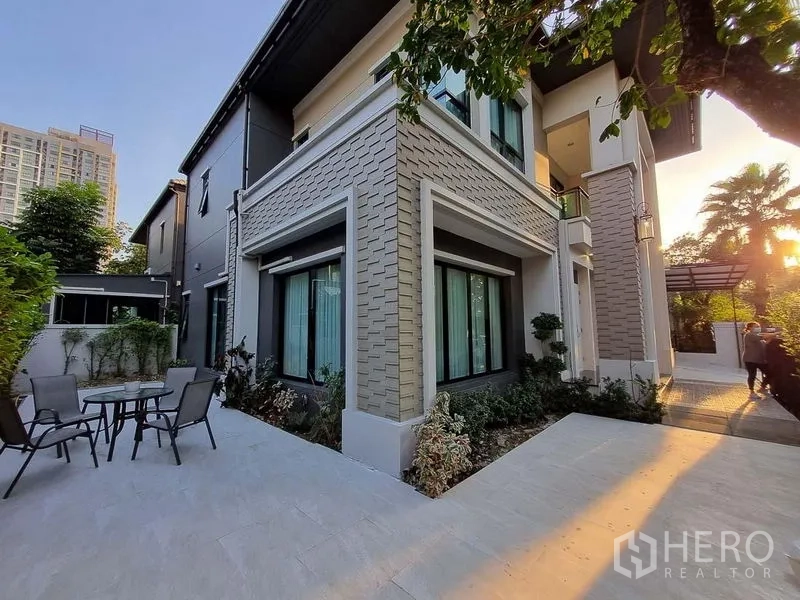 Detached House for rent in Bang Khae, Bangkok - Side of the house with patio, landscaped beds, and large windows at sunset.