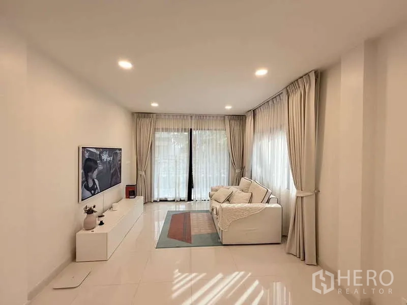บ้าน ขายในBang Phli, Samut Prakan - ห้องนั่งเล่นกว้างพร้อมโซฟา ผนังทีวี และผ้าม่านสูงจรดพื้นเปิดสู่สวน