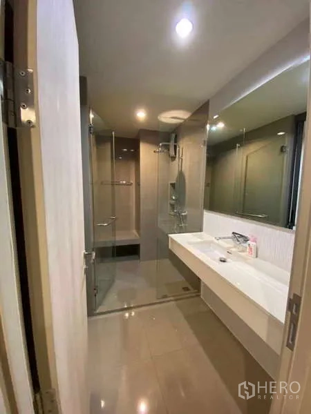 Condominium for rent or sale in Khlong Toei, Bangkok - Modern bathroom with long vanity, large mirror and glass‑enclosed rain shower.