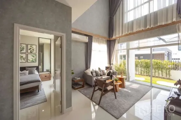 Detached House for sale in Lam Luk Ka, Pathum Thani - Bright living room with double-height ceiling, floor-to-ceiling glass and adjacent bedroom suite.