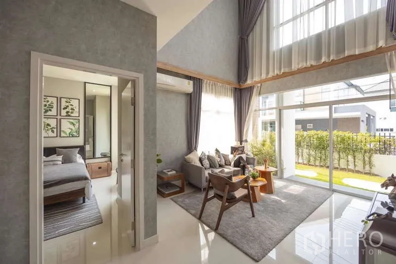 Detached House for sale in Lam Luk Ka, Pathum Thani - Bright living room with double-height ceiling, floor-to-ceiling glass and adjacent bedroom suite.