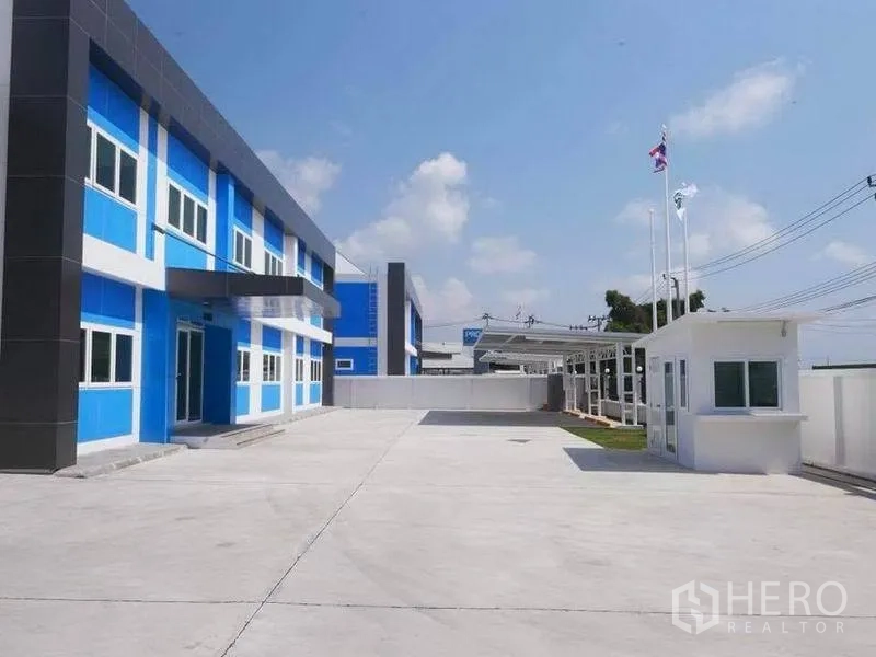 Factory for rent in Bang Bo, Samut Prakan - Wide driveway and parking apron with guardhouse, flags and blue–grey office façade within a secure walled compound.
