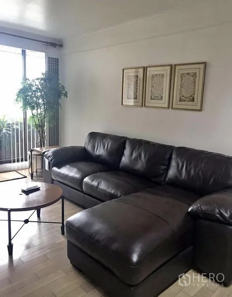 Condominium for rent in Khlong Toei, Bangkok - Living room with dark leather sectional sofa, round coffee table and balcony access through sliding glass doors.