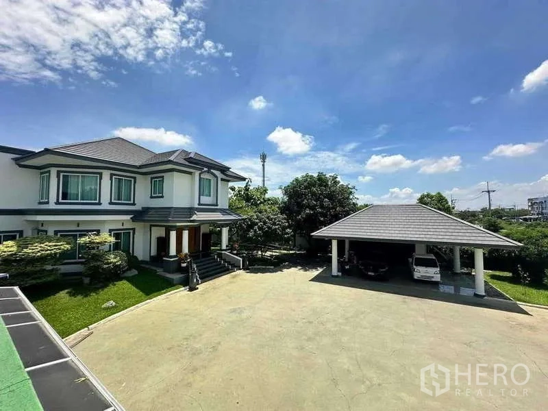 Detached House for sale in Khlong Luang, Pathum Thani - Expansive driveway with the main two-storey house and a covered carport amid green lawns under a blue sky.