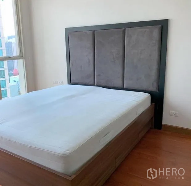 Condominium for rent in Pathum Wan, Bangkok - Bedroom with large upholstered headboard, city-view window, and warm wood flooring in a Pathum Wan condo.
