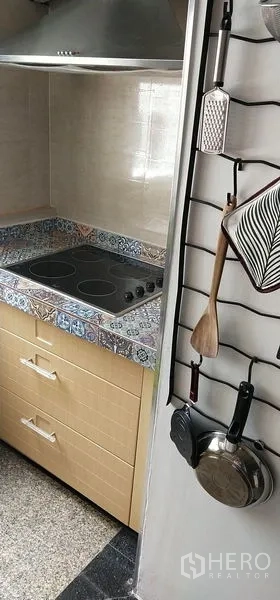 Condominium for sale in Watthana, Bangkok - Cooking corner with electric cooktop, patterned tile countertop and hanging kitchen utensils.