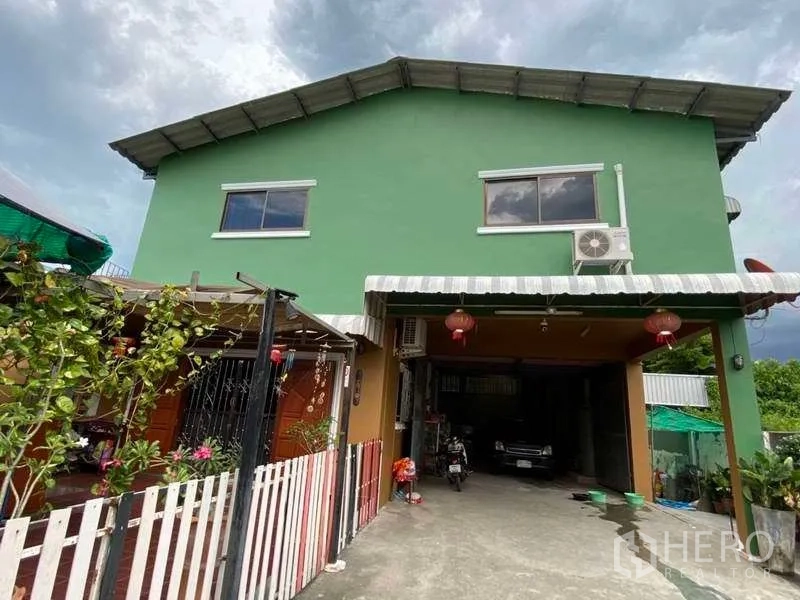 货栈 出租或出售在Pak Kret, Nonthaburi - 车道通向绿色两层仓库的有顶装卸区，篱笆旁有绿植。