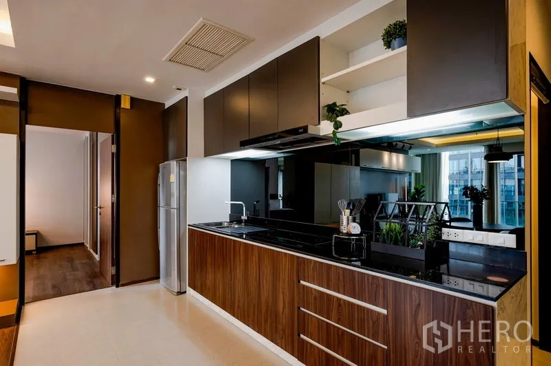 Condominium for sale in Khlong Toei, Bangkok - Modern linear kitchen with wood cabinets, black countertop, mirrored backsplash, and stainless refrigerator.