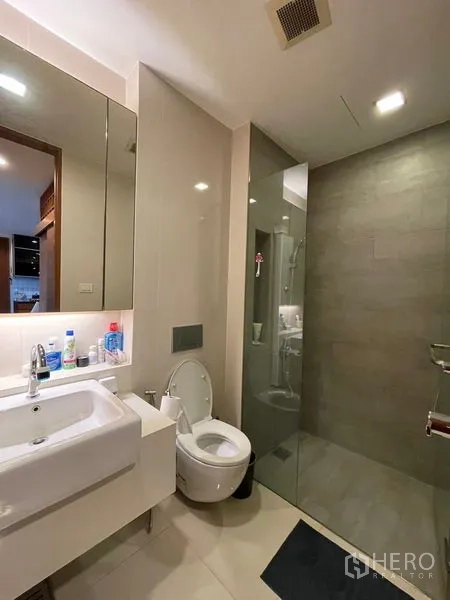 Condominium for sale in Khlong Toei, Bangkok - Modern bathroom with wall-mounted sink, toilet and a glass-enclosed shower in neutral tiles.