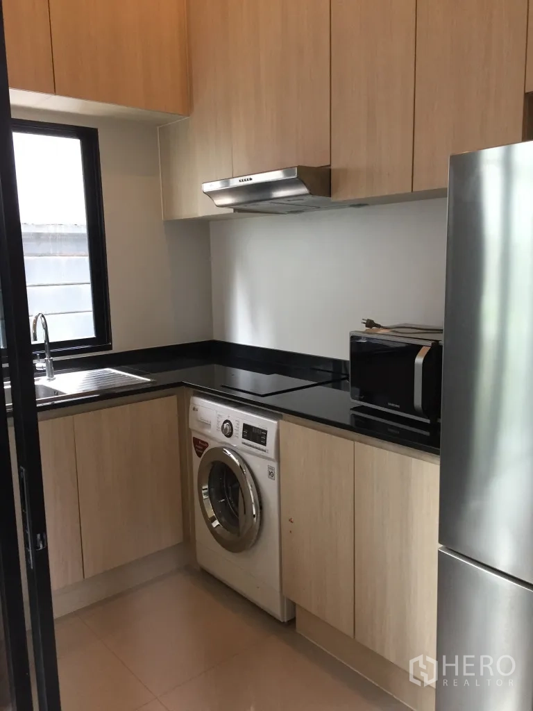 Townhouse for rent in Suan Luang, Bangkok - Enclosed modern kitchen with light wood cabinets, black countertops, window, sink, hood, fridge and washer.